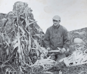 black and white photo of a man kneeling with corn and cornstalks