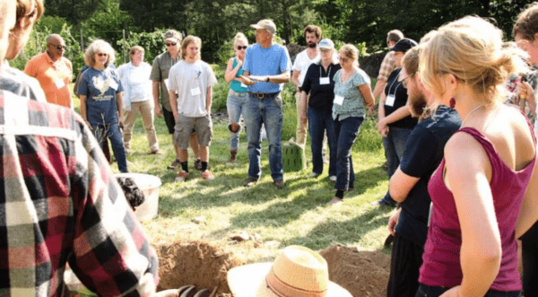 A group of students gather around an instructor on a farm, on a bright and sunny day.